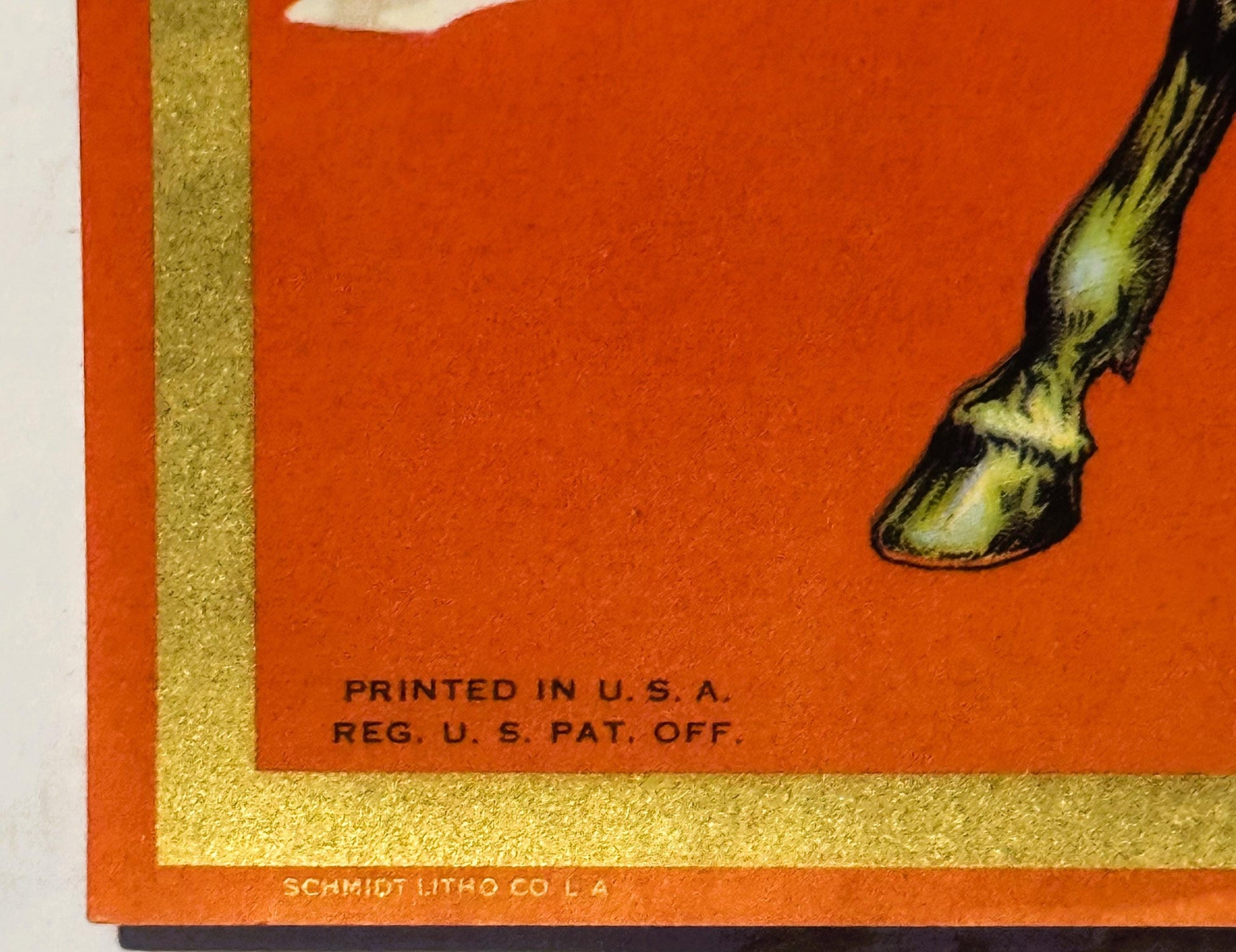 Close-up of vintage orange crate label corner with gold border showing horse's hoof, "PRINTED IN U.S.A." and "SCHMIDT LITHO C