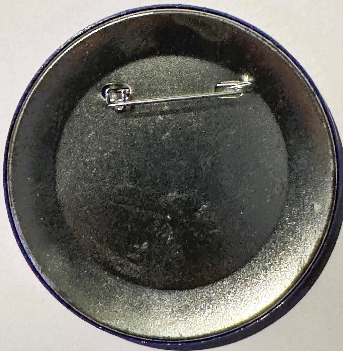 Back of a round metal pin with safety clasp, gray powdery surface, navy blue rim, photographed from above