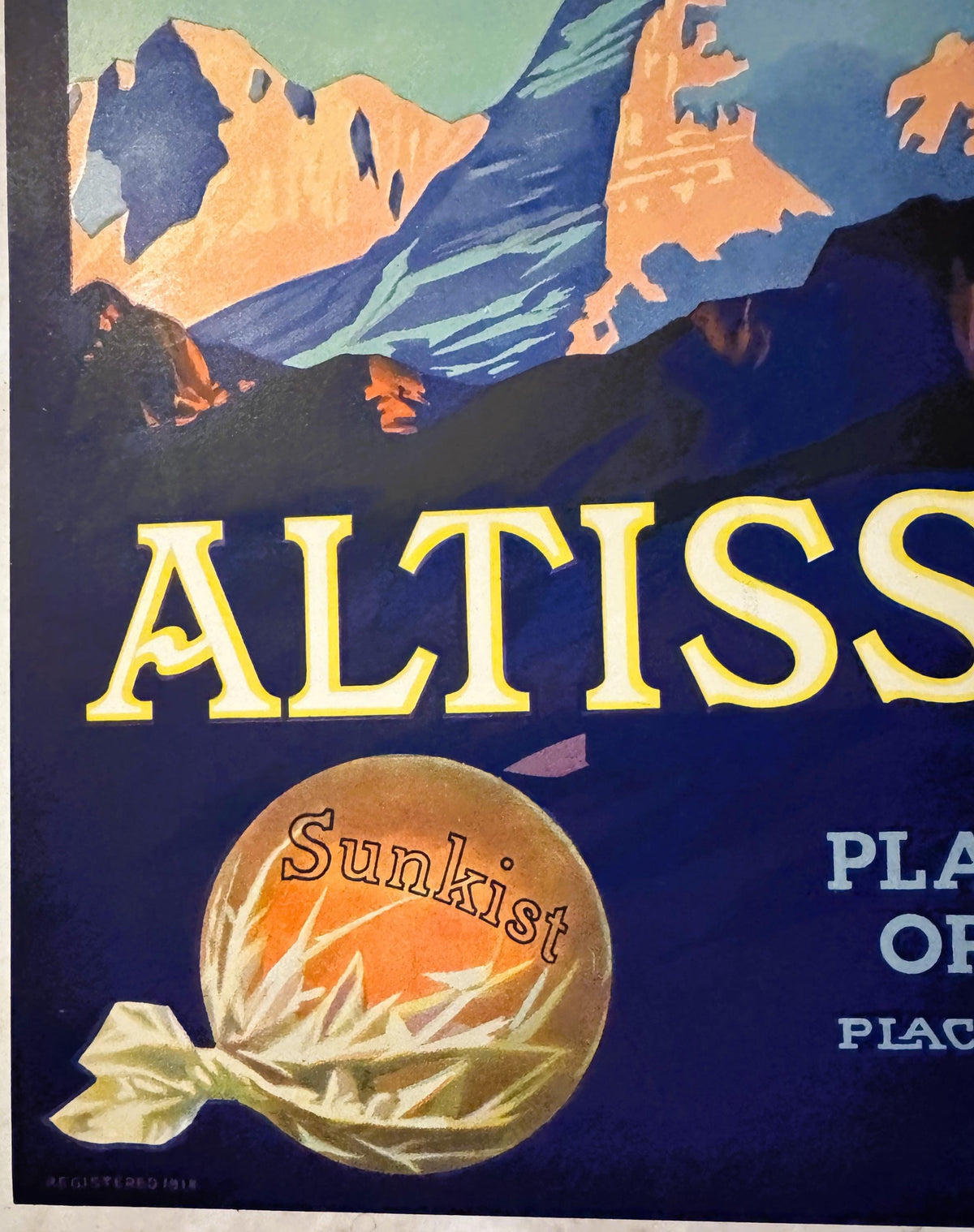 Close-up of the Altissimo crate label's lower left: a Sunkist orange wrapped in white tissue twisted at the end, with Registered 1918 in tiny type beneath it