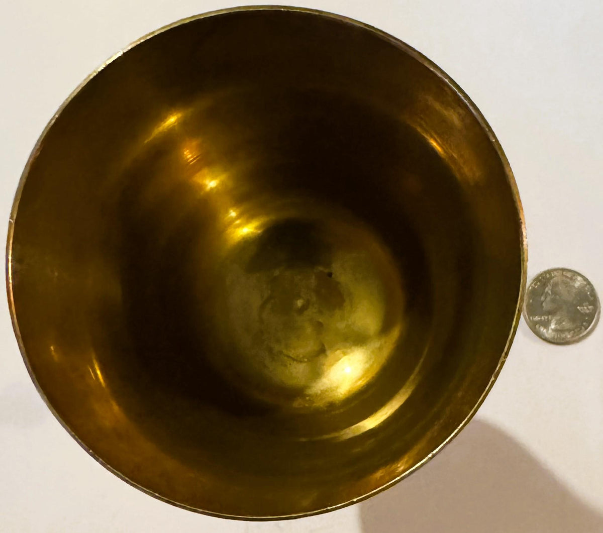 Overhead view of a brass goblet with golden-brown patina and etched details inside, shown next to a quarter for scale