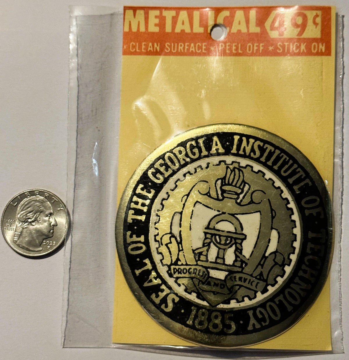 Vintage metallic Georgia Institute of Technology decal on yellow backing with quarter for scale, next to clear protective sle