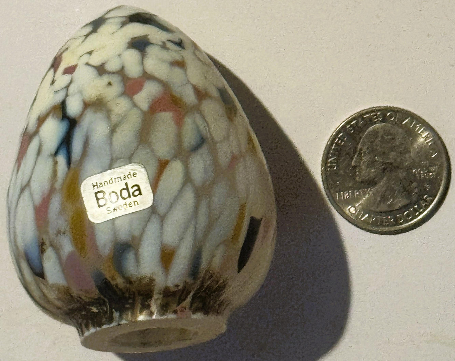 Handmade ceramic egg with cream, rust, and blue speckled glaze next to a coin for scale, labeled "Boda Sweden