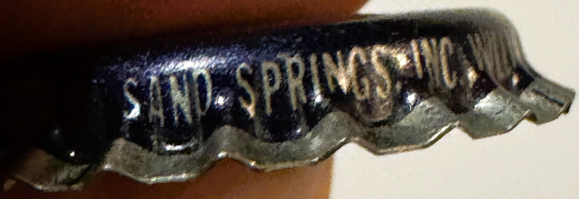 Close-up of dark blue vintage bottle cap with embossed "SAND SPRINGS" text and metallic ridged edge, held against brown fabri