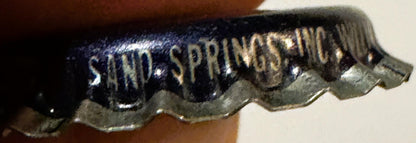 Close-up of dark blue vintage bottle cap with embossed "SAND SPRINGS" text and metallic ridged edge, held against brown fabri