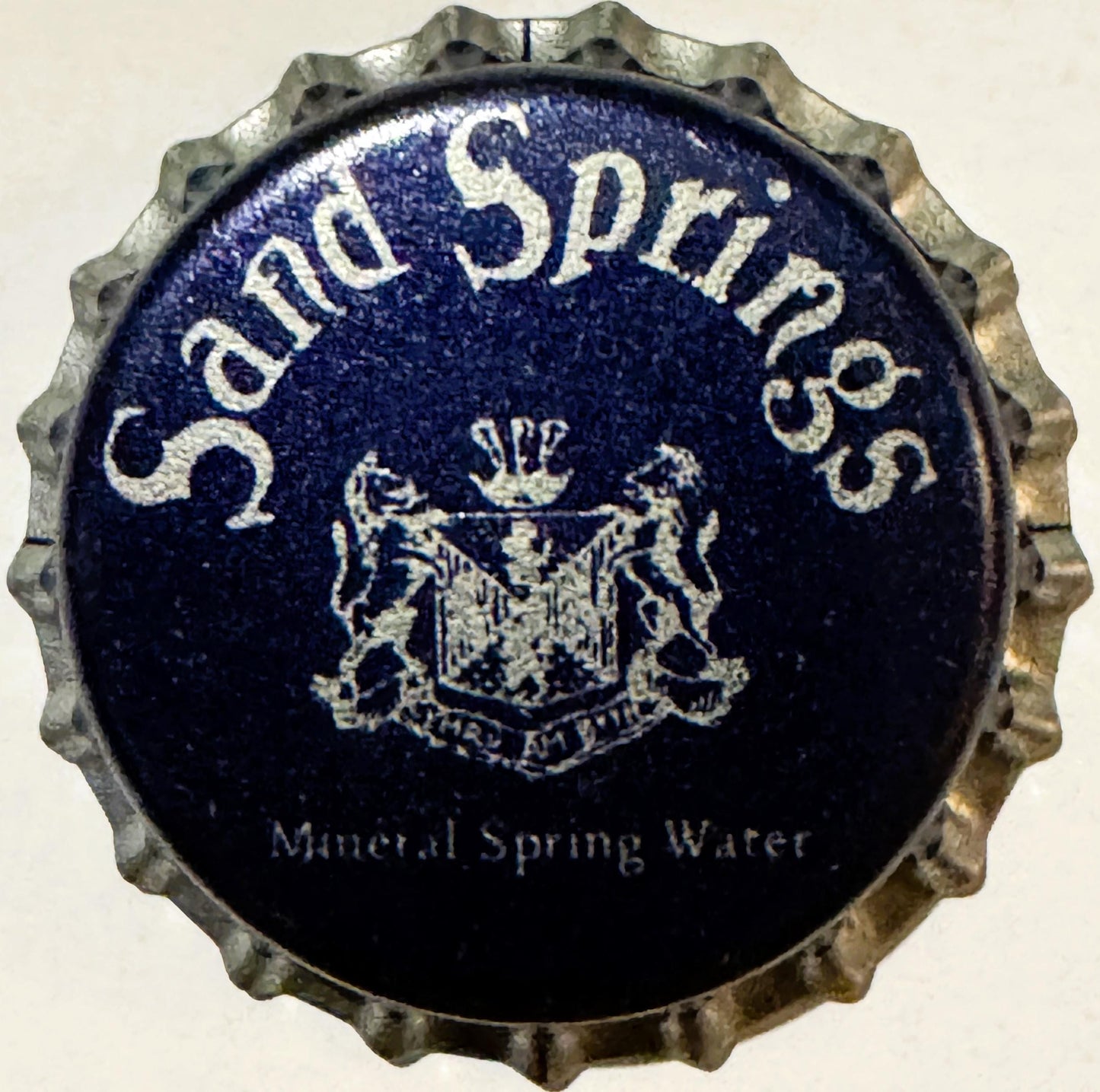 Vintage bottle cap with corrugated metal rim and dark blue center printed with "Sand Springs" text, crest emblem, and "Minera