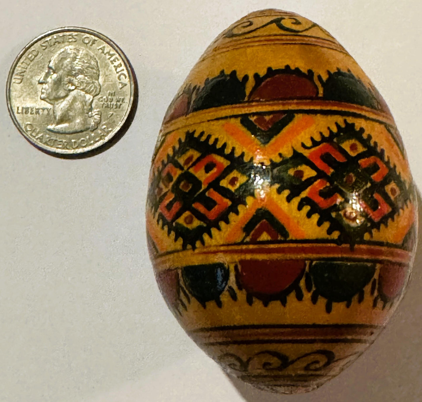 Wooden egg with hand-painted geometric patterns in red, orange, black and tan, displayed next to a quarter for scale