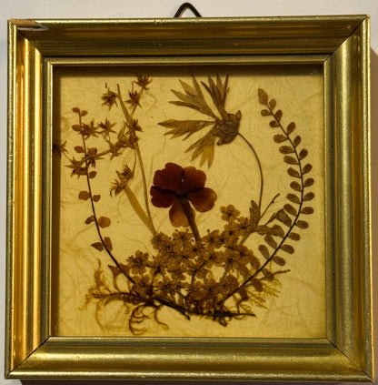 Small square pressed-flower picture in a gilt frame: a dark red bloom at centre, a fern frond and star-shaped sprigs framing it, yellow-tan dried blossoms and moss along the bottom, on cream textured paper. Front view, hanger visible at top.