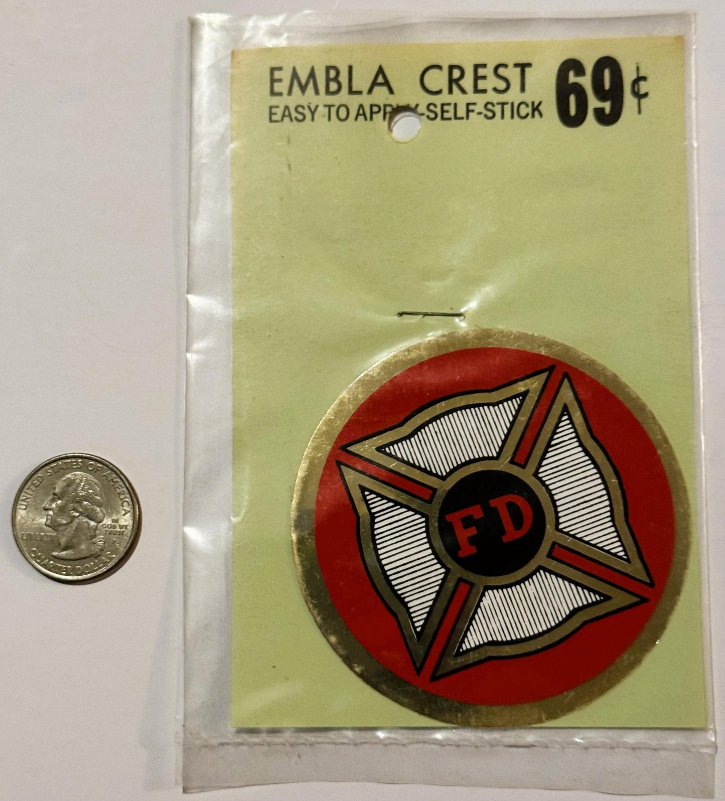 Vintage self-stick emblem crest in original packaging showing red Maltese cross with "FD" center, next to quarter for scale