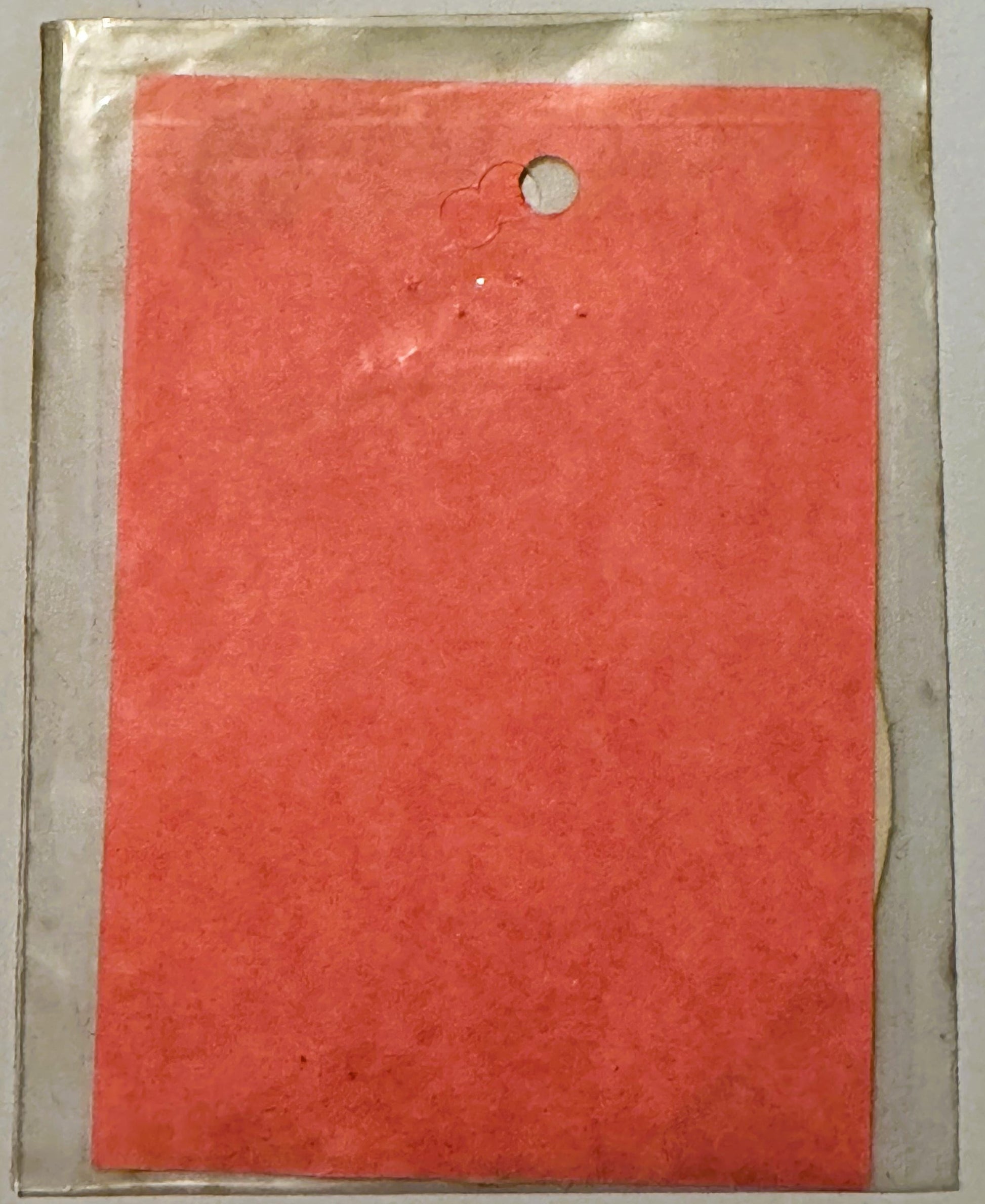 Bright coral-red rectangular decal or sticker in clear plastic sleeve, showing wear marks and a small hole near top center