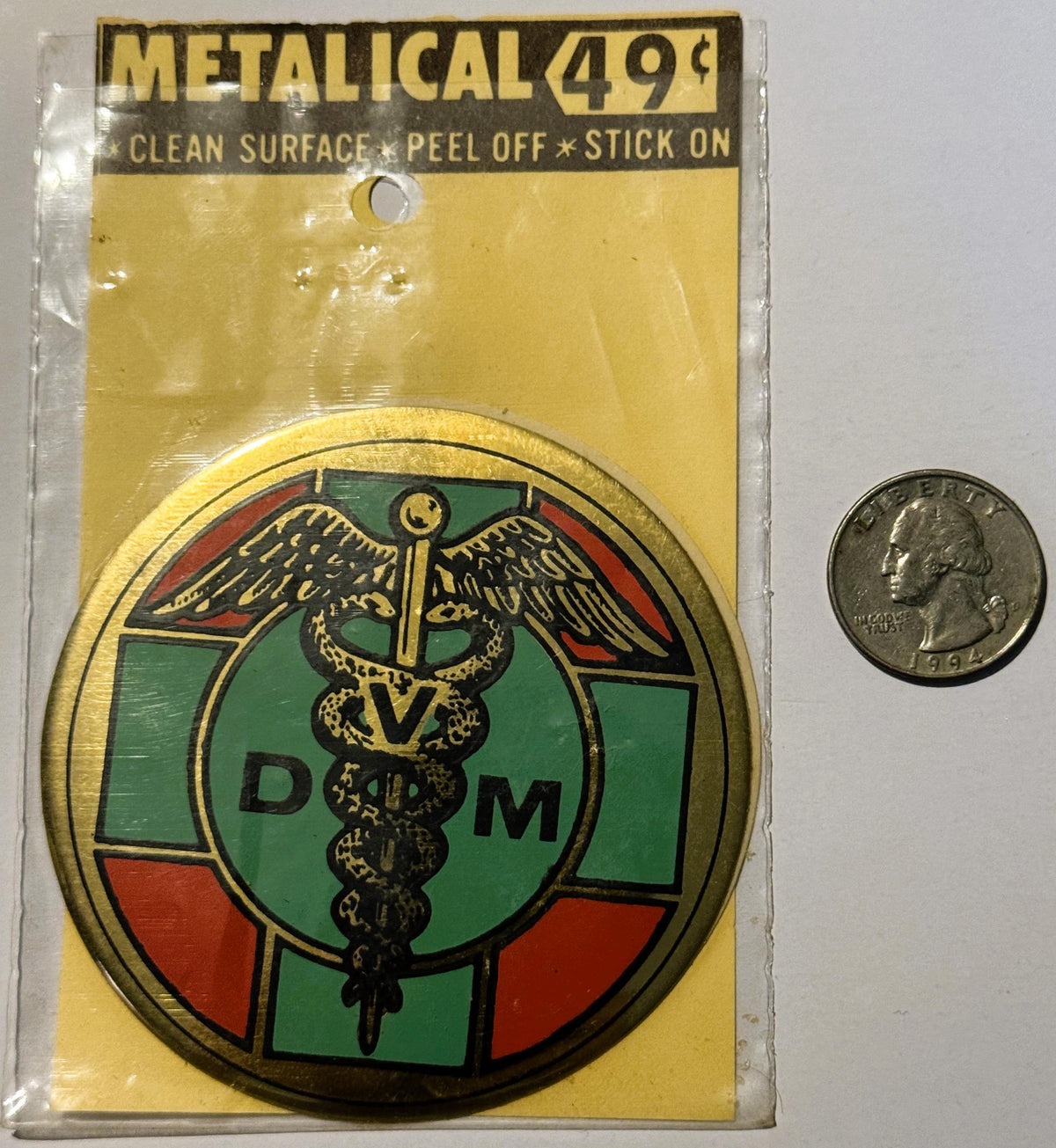 Vintage metallic decal sticker with DVM caduceus medical symbol in green, red, and gold, next to a quarter for scale