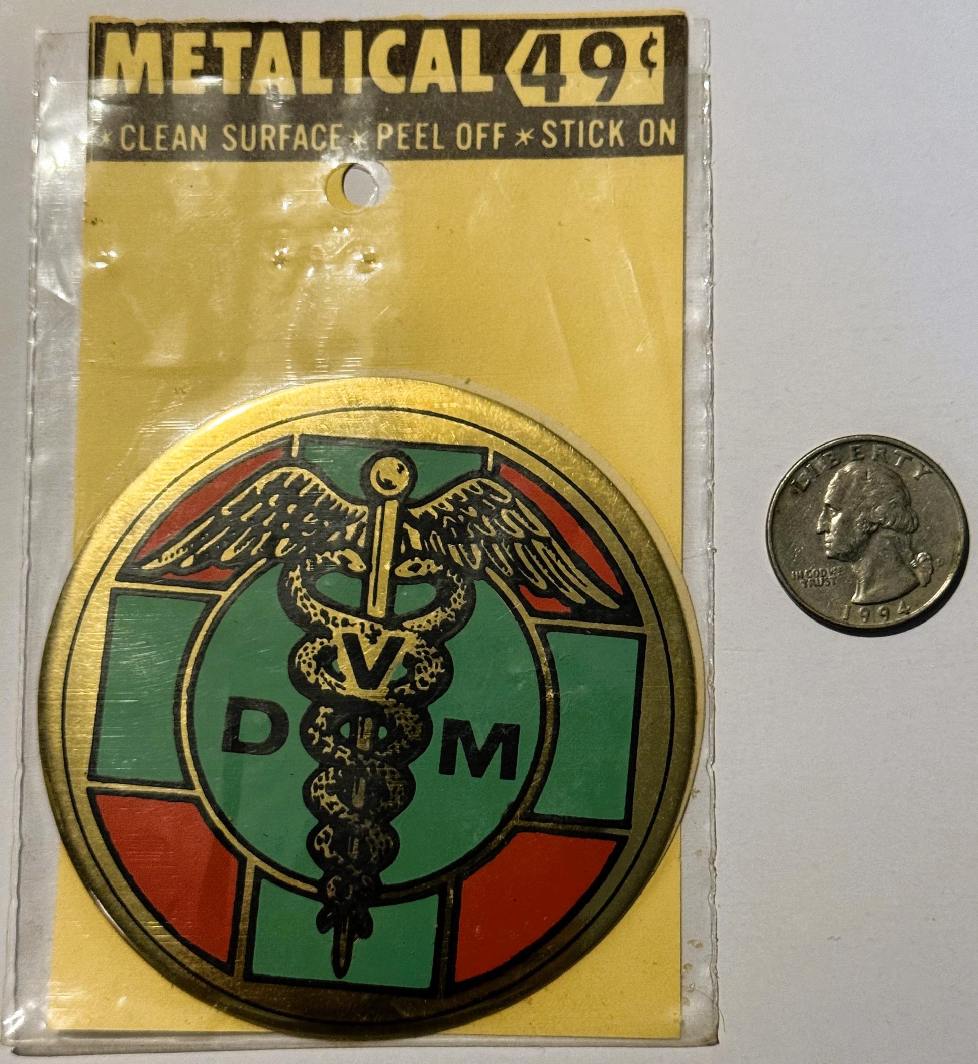Vintage metallic decal sticker with DVM caduceus medical symbol in green, red, and gold, next to a quarter for scale
