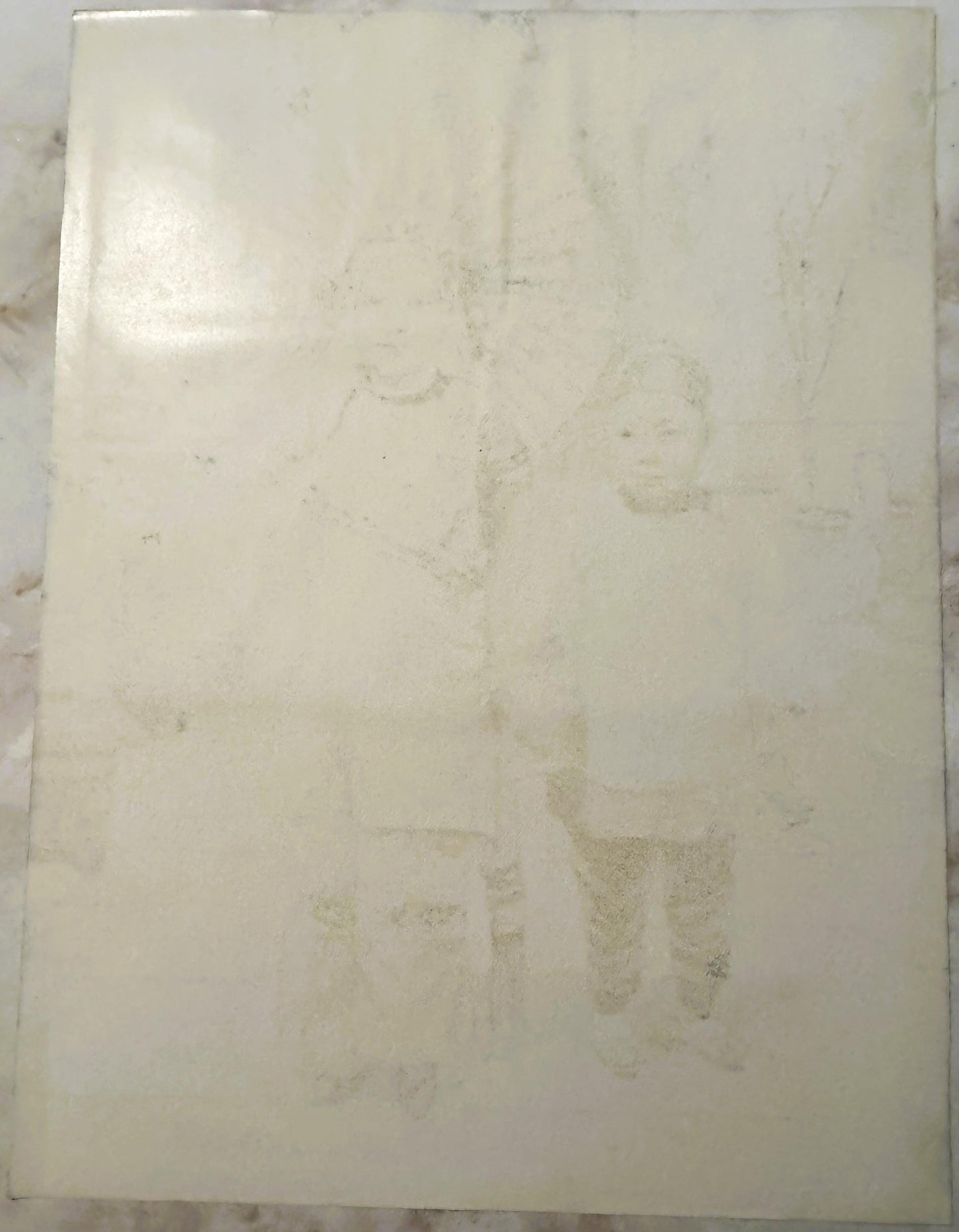 Cream-colored antique print with faded imagery of figures, visible wear and staining throughout, displayed flat against marbl