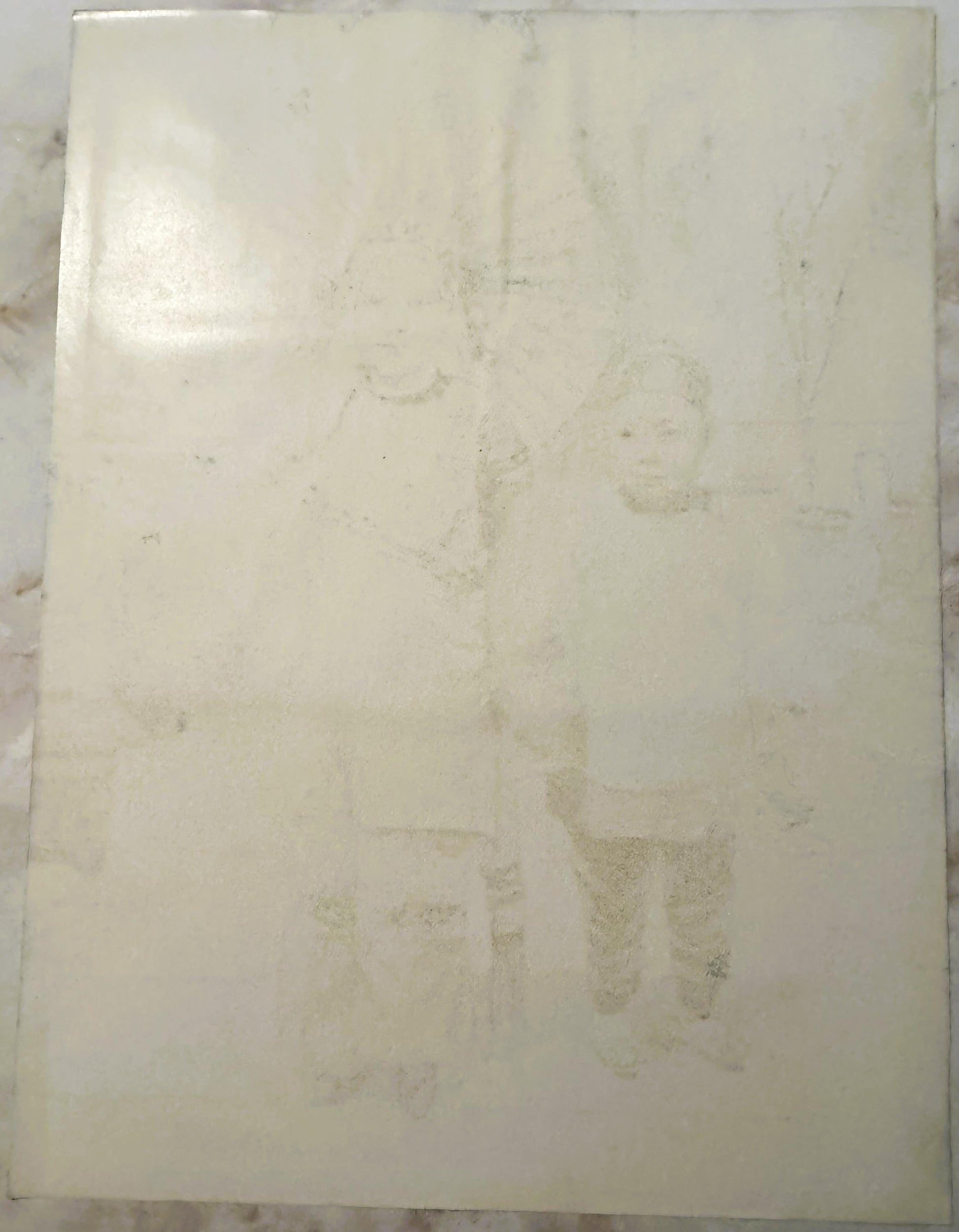 Cream-colored antique print with faded imagery of figures, visible wear and staining throughout, displayed flat against marbl