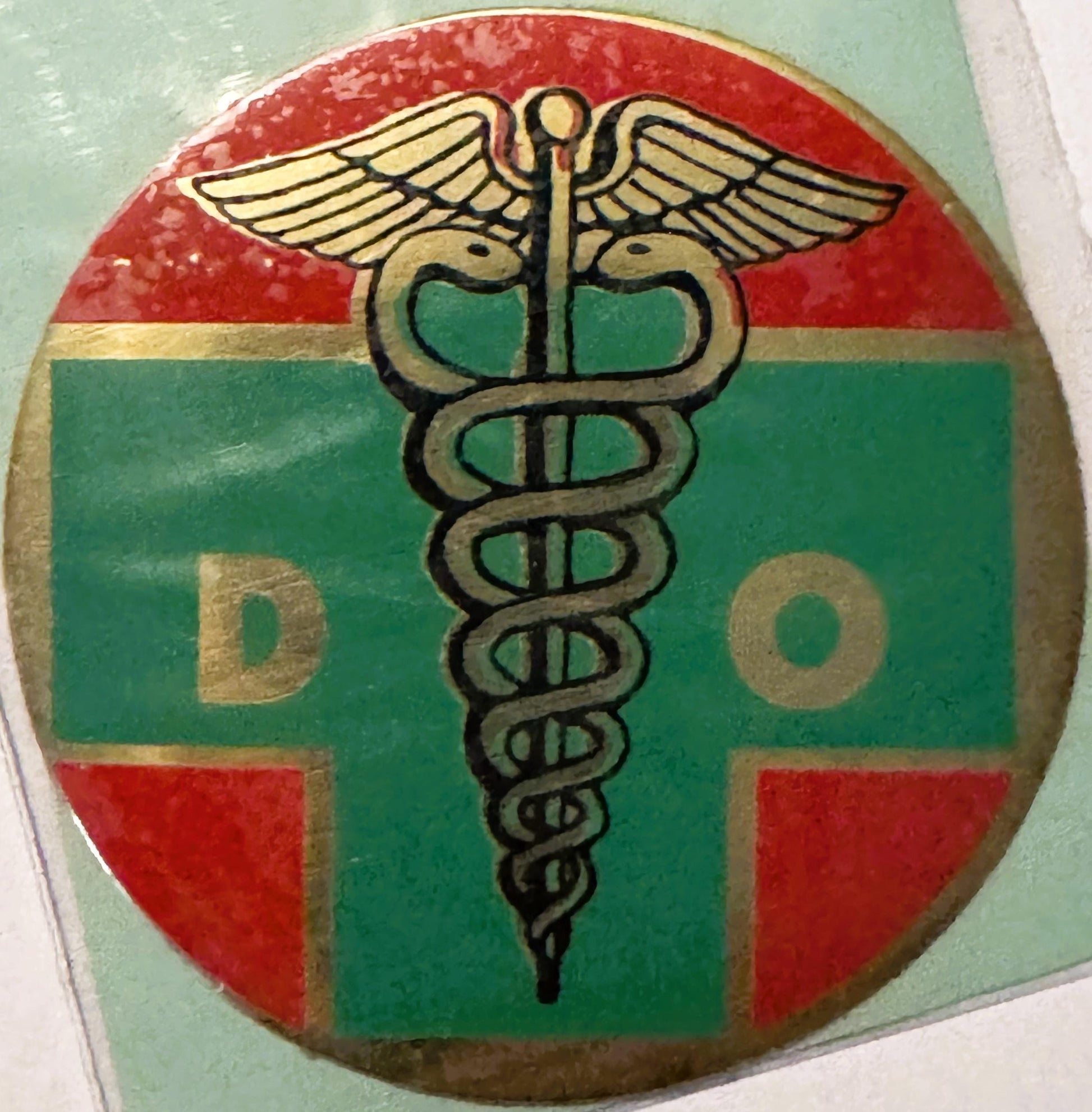 Circular vintage decal with red and green quarters featuring a caduceus medical symbol and letters "D" and "O" in gold and bl