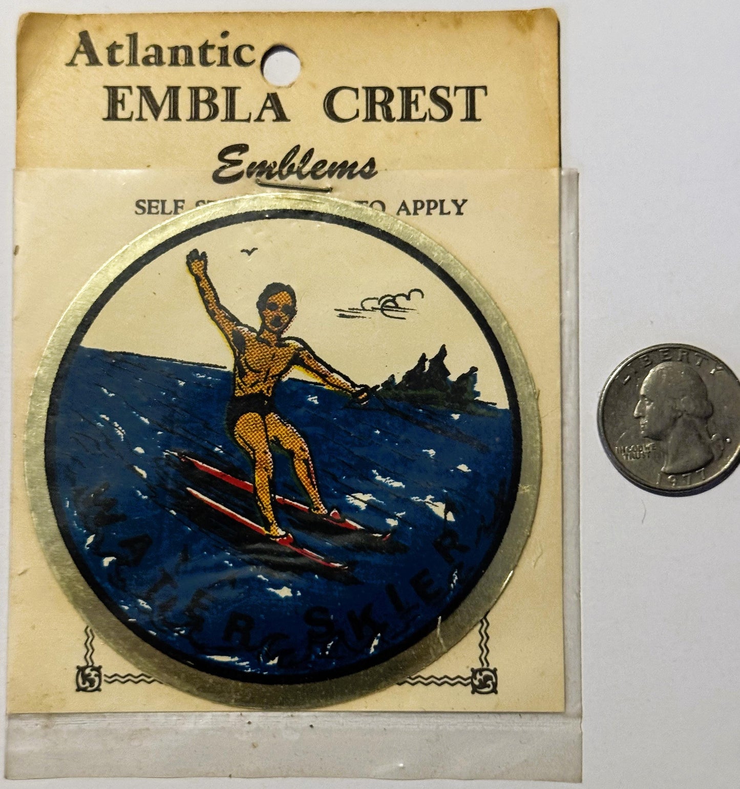 Vintage Atlantic Embla Crest water skier decal on original packaging with quarter for scale, featuring gold figure on red ski