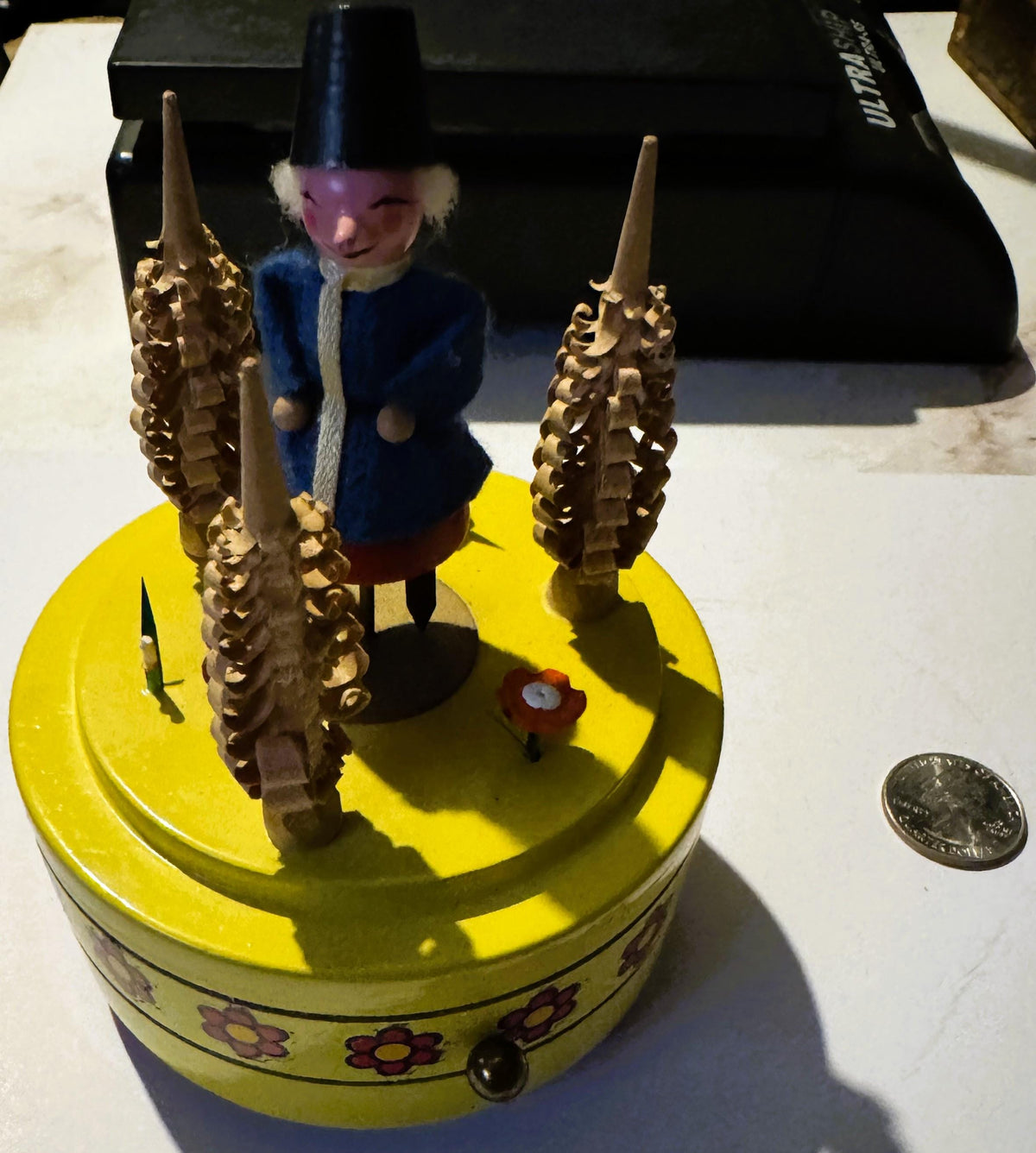 Yellow vintage music box with ornate brass spires and dancing folk figure in blue coat, displayed next to a coin for scale