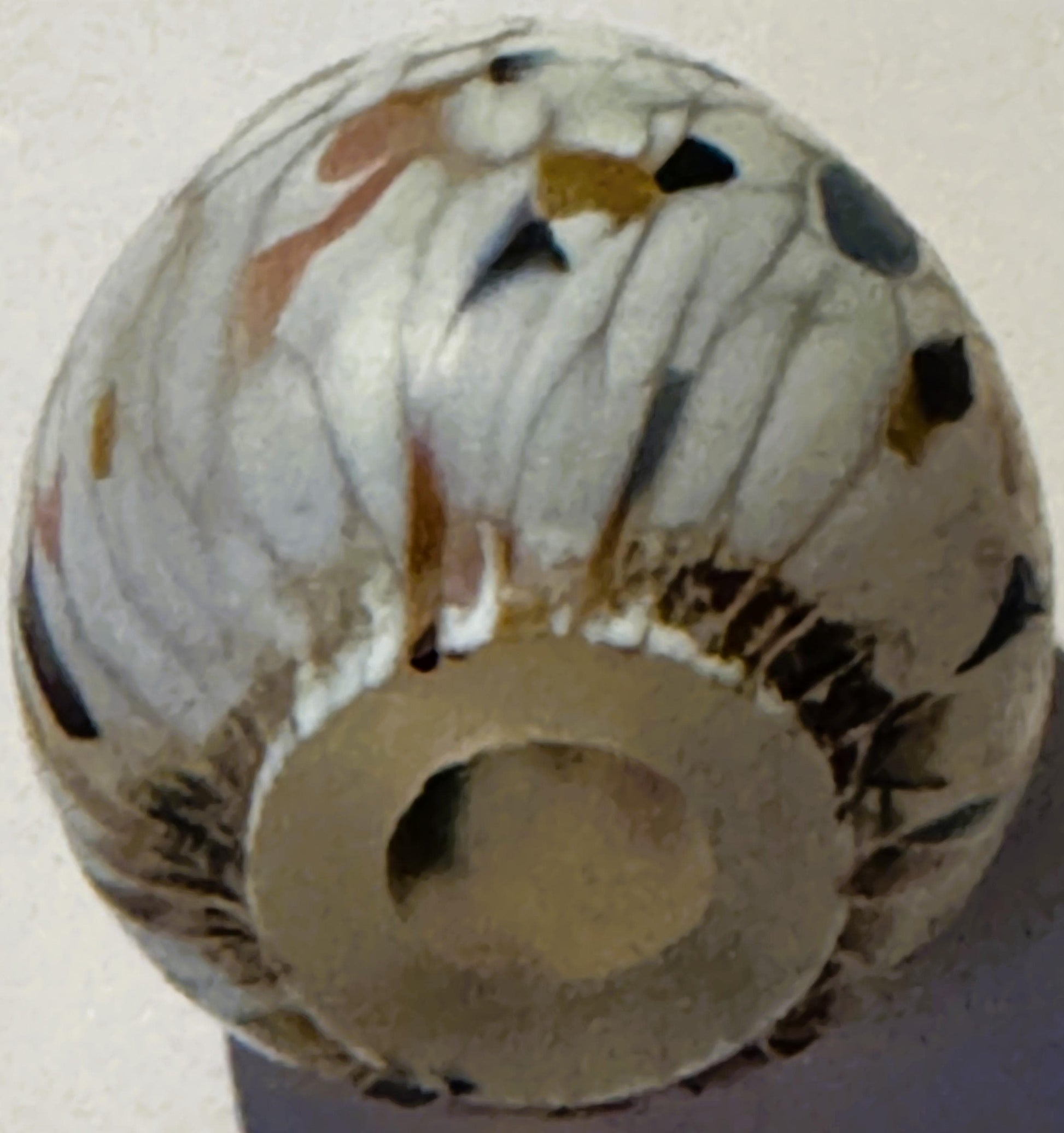 Cream-colored glass egg with brown, black, and gold spattered confetti pattern, viewed from above against a light background