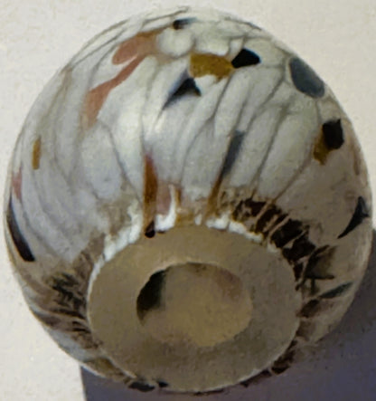 Cream-colored glass egg with brown, black, and gold spattered confetti pattern, viewed from above against a light background