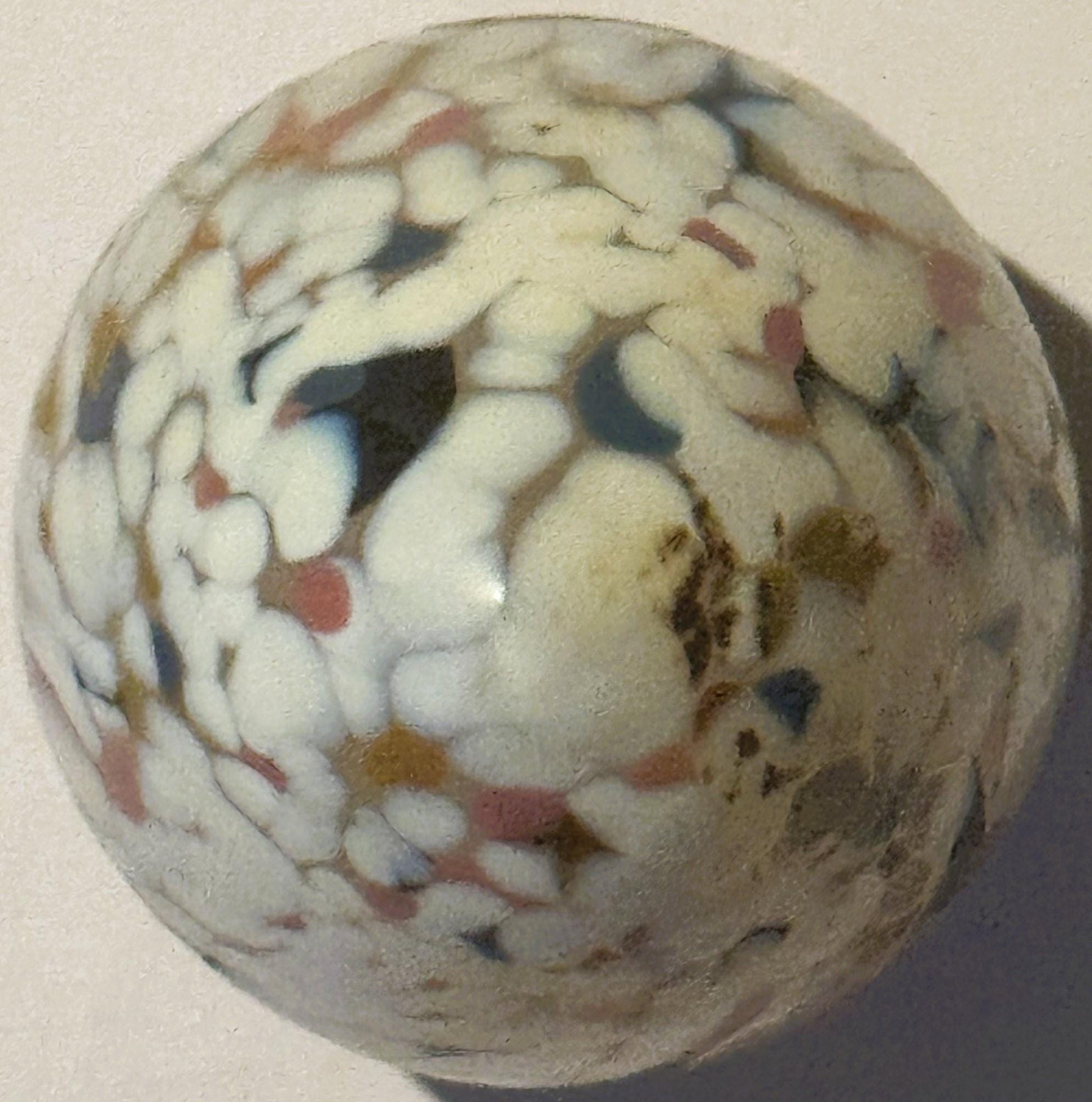 Rounded cream-colored glass egg with multicolored confetti specks in red, blue, black, and gold, photographed against a beige