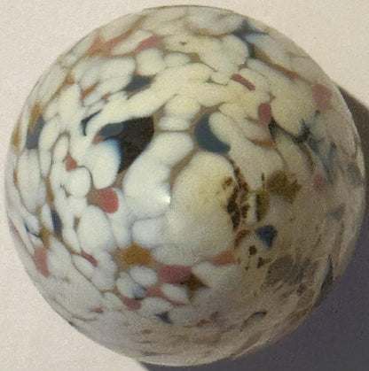 Rounded cream-colored glass egg with multicolored confetti specks in red, blue, black, and gold, photographed against a beige