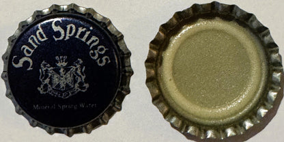 Vintage bottle cap with ridged metal edge, dark blue front showing "Sand Springs" text and coat of arms emblem, plain metalli