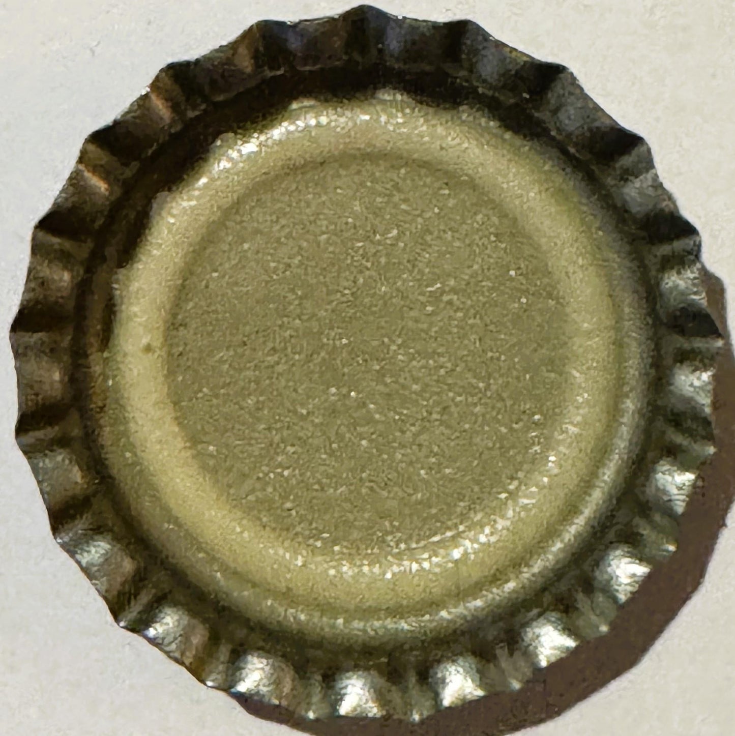 Unused metal bottle cap with ridged edge and embossed center, tan and dark brown patina, viewed from above against white back
