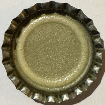 Unused metal bottle cap with ridged edge and embossed center, tan and dark brown patina, viewed from above against white back