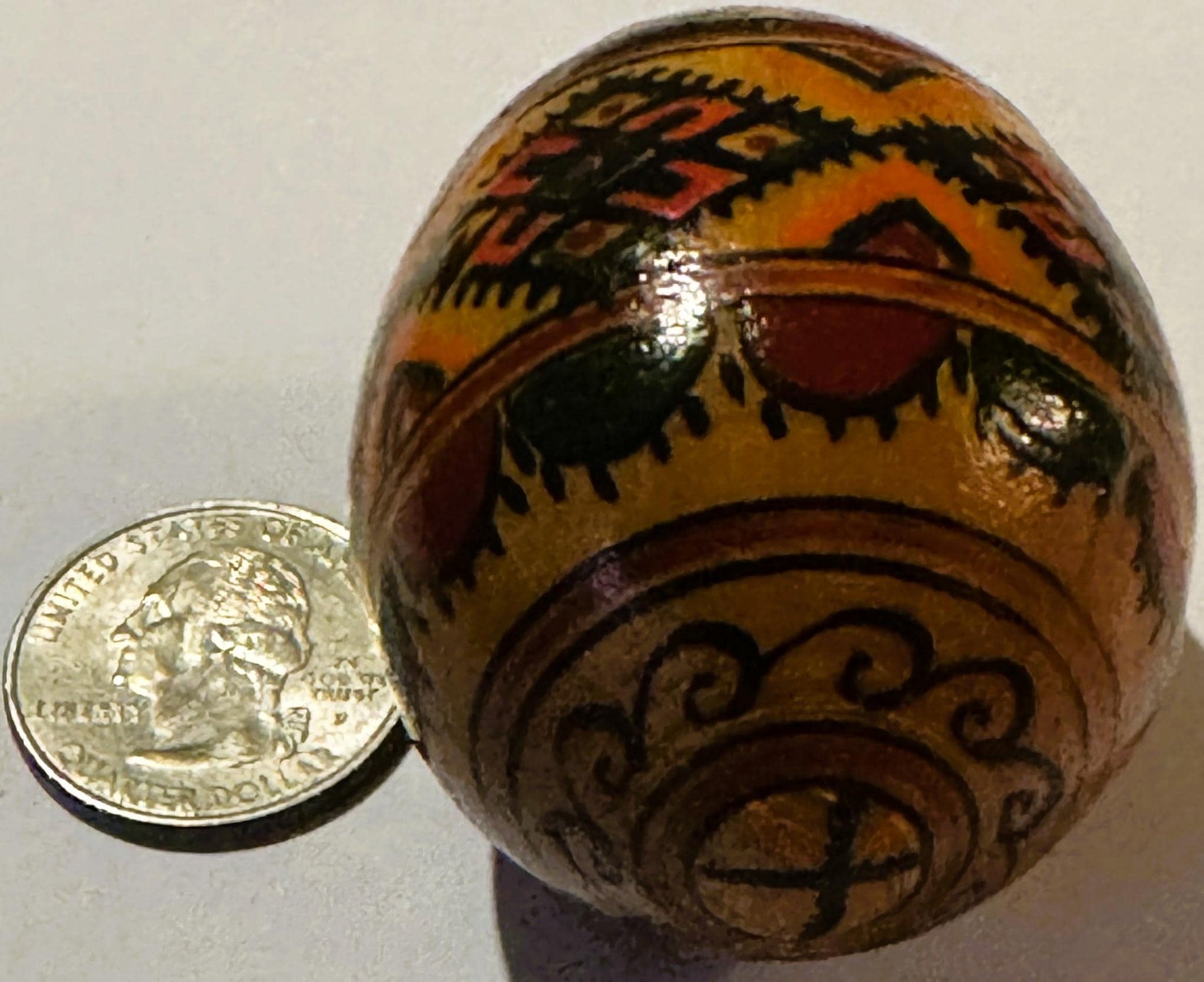Wooden egg with hand-painted geometric patterns in orange, black, and red, displayed next to a US quarter for scale.