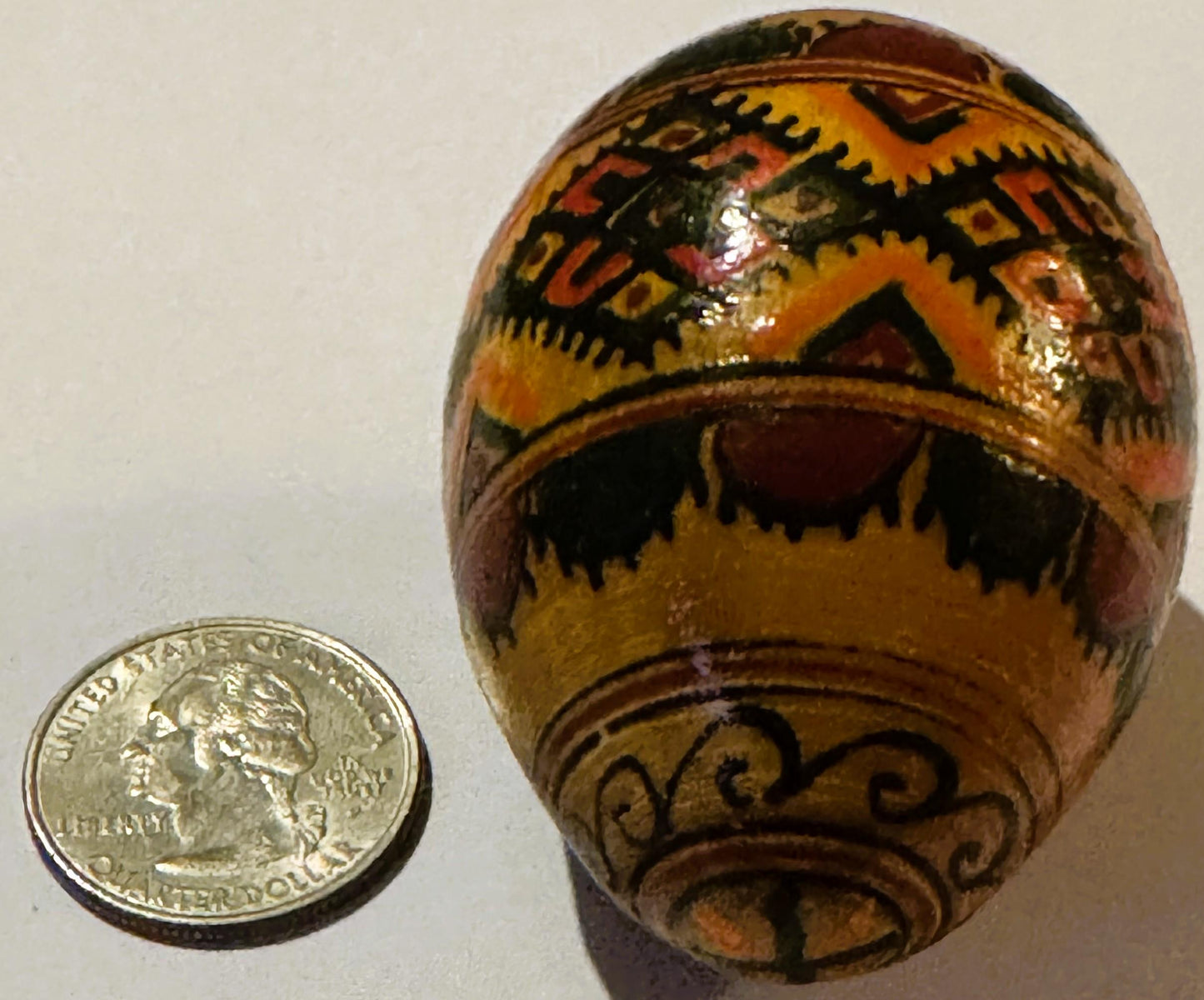 Hand-painted wooden egg with geometric patterns in orange, yellow, black and brown, displayed next to a US quarter for scale