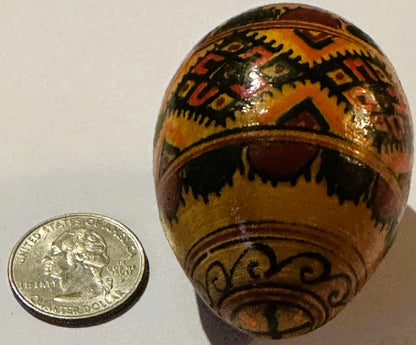 Hand-painted wooden egg with geometric patterns in orange, yellow, black and brown, displayed next to a US quarter for scale