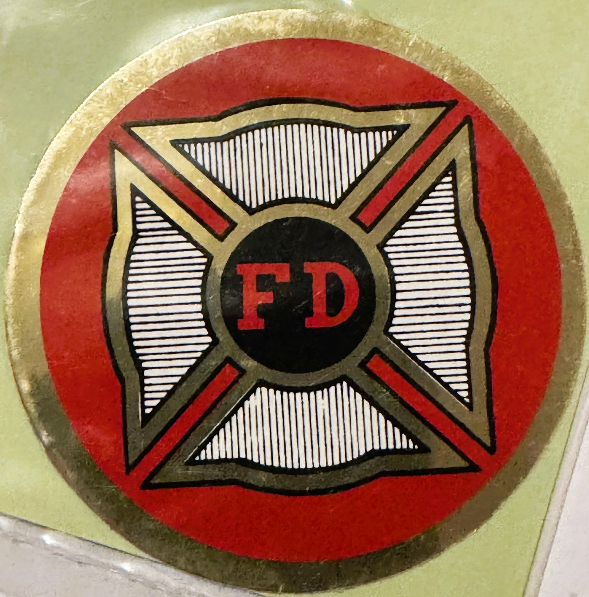 Circular red and gold vintage FD firefighter decal with Maltese cross design and black center circle, mounted on pale green s
