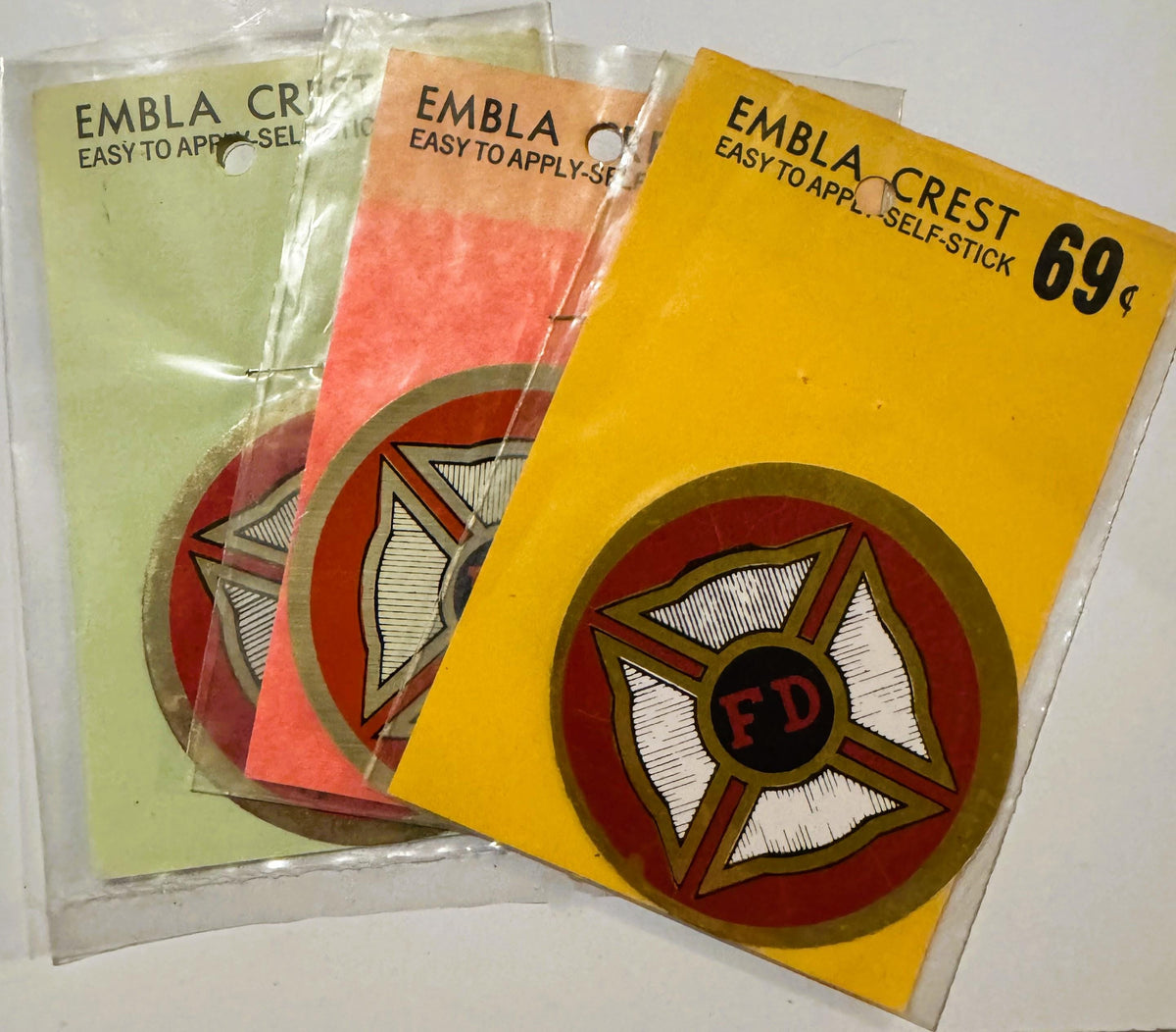 Three vintage Embla Crest self-stick decals in yellow, coral, and pale green with red Maltese cross and "FD" center emblems, 
