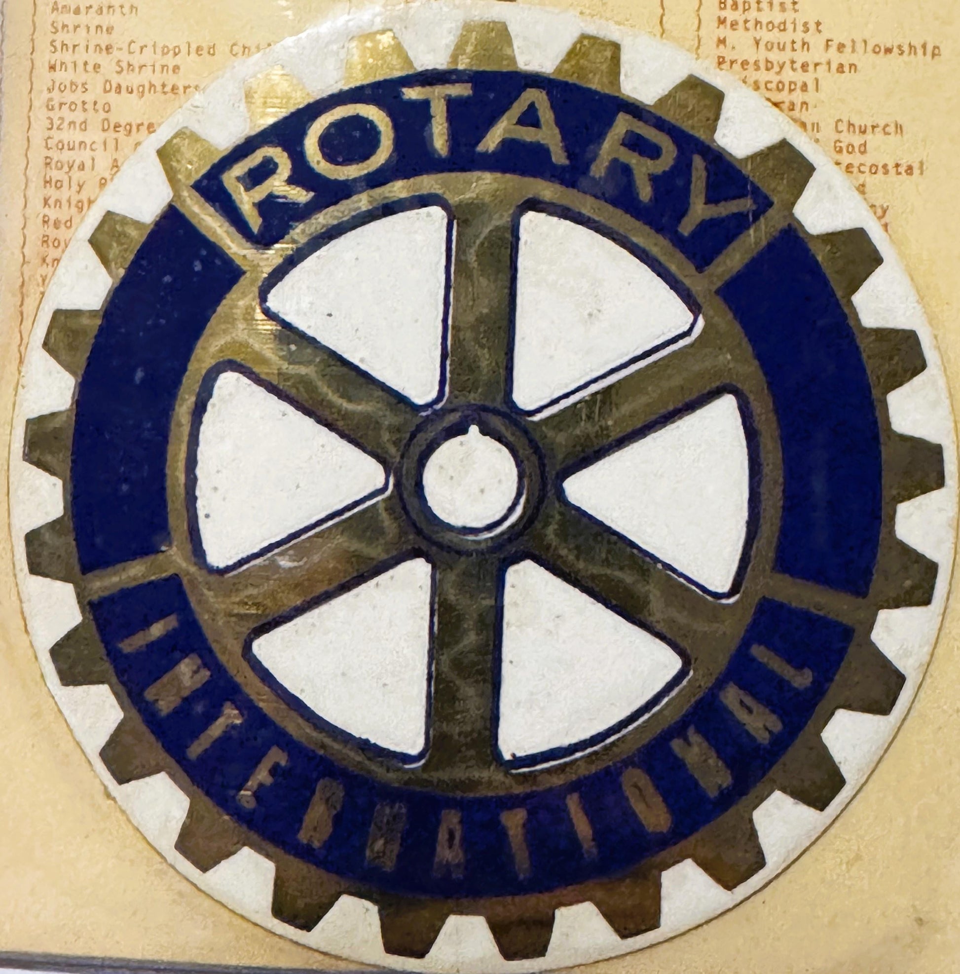 Rotary International emblem decal with gear-shaped border, white cross on bronze center, dark blue ring, mounted on aged pape