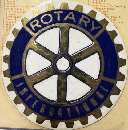 Rotary International emblem decal with gear-shaped border, white cross on bronze center, dark blue ring, mounted on aged pape