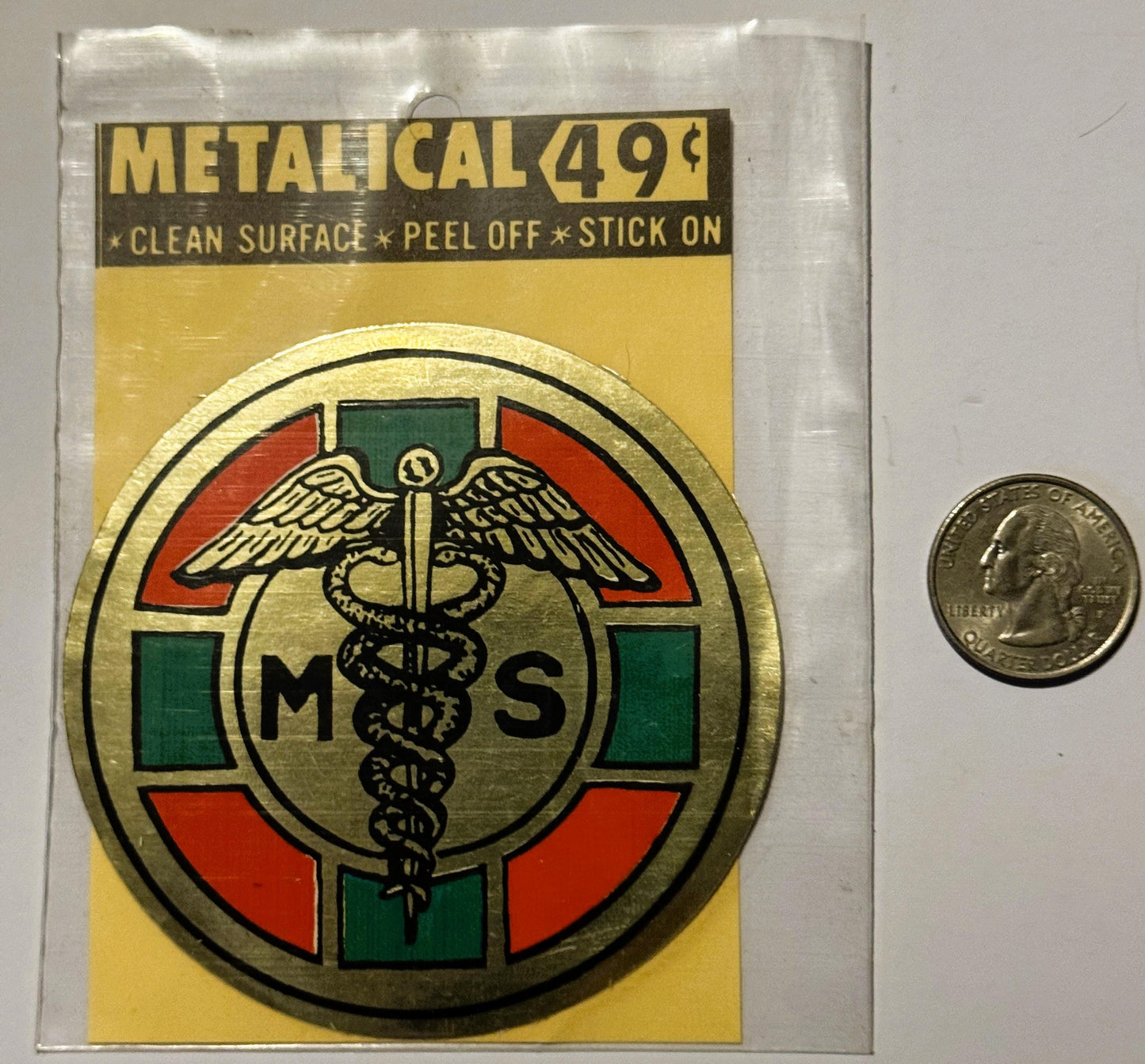 Metallic medical caduceus sticker in original packaging with quarter for scale, showing winged staff symbol with "MS" letters