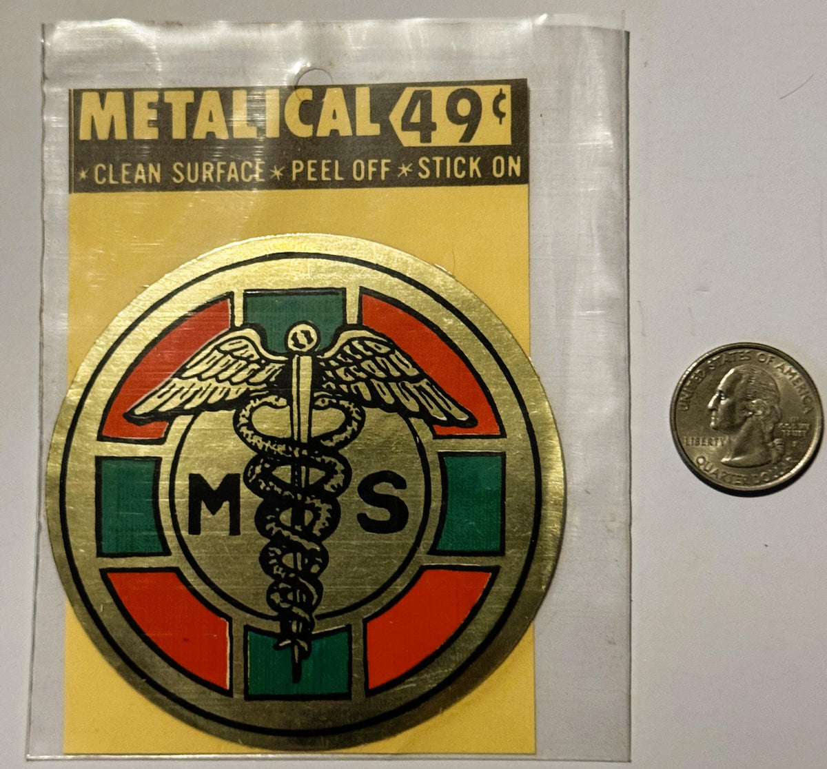 Metallic medical caduceus sticker in original packaging with quarter for scale, showing winged staff symbol with "MS" letters