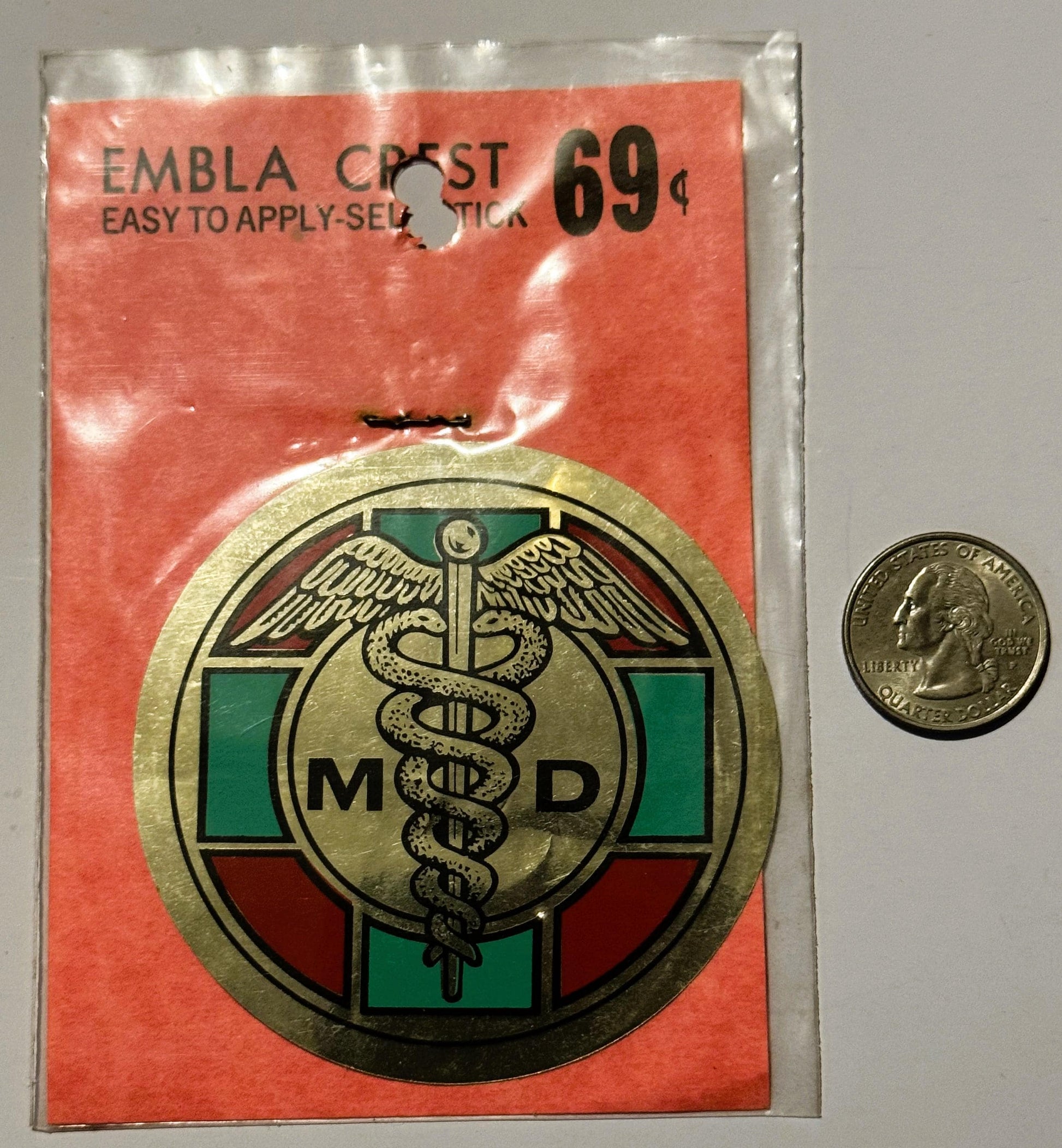 Vintage medical emblem decal in plastic packaging with quarter coin for scale, showing caduceus symbol with "MD" text and red