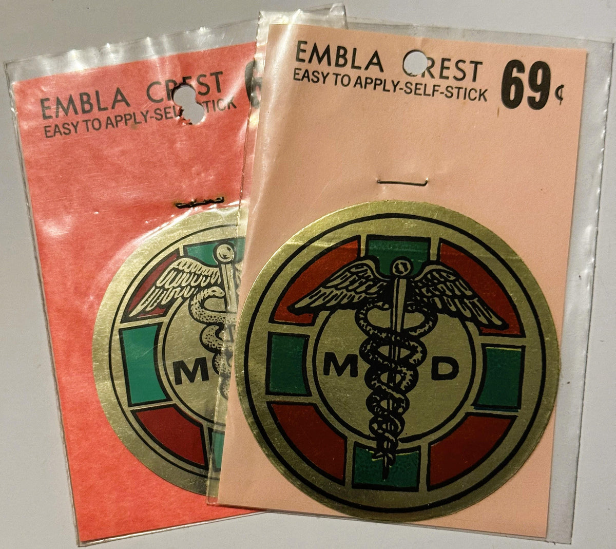 Two vintage self-stick medical decals in original packaging: coral-colored and tan, each featuring an MD caduceus emblem with