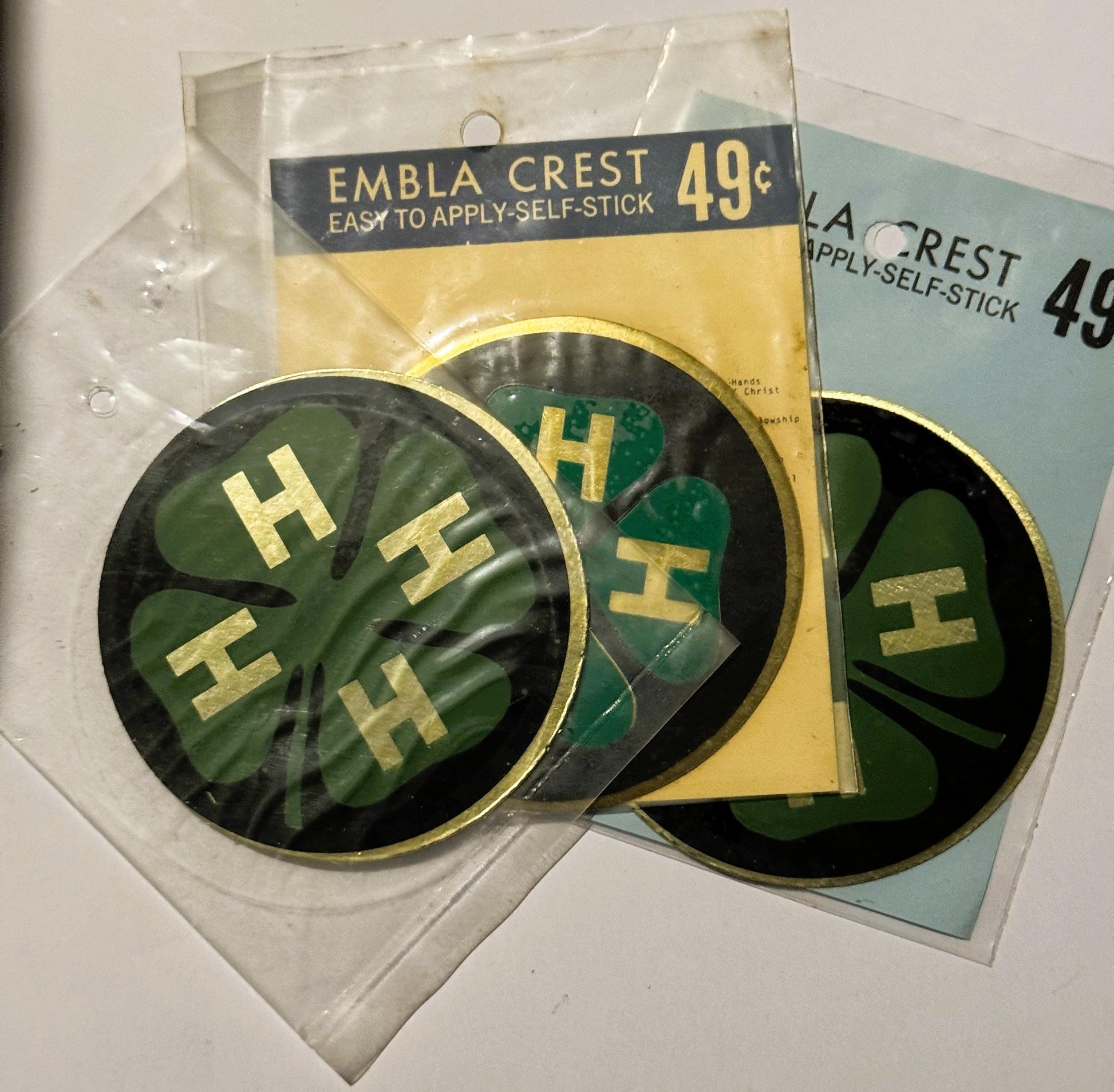 Two circular green and gold 4H emblem decals in original packaging labeled "Embla Crest Easy to Apply Self-Stick 49¢" display