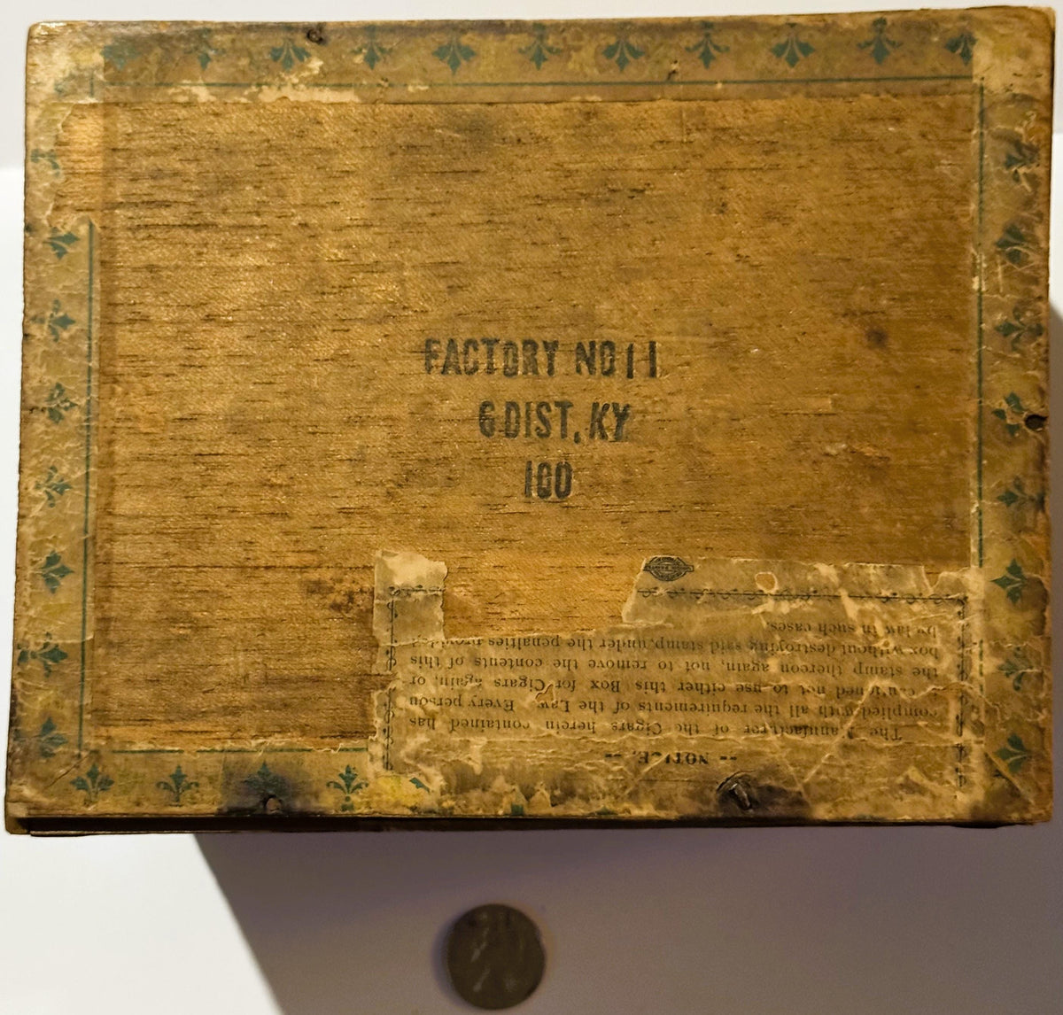 Aged wooden cigar box lid with decorative fleur-de-lis border, stamped "Factory No. 11, 6 Dist. KY, 100," penny shown for sca