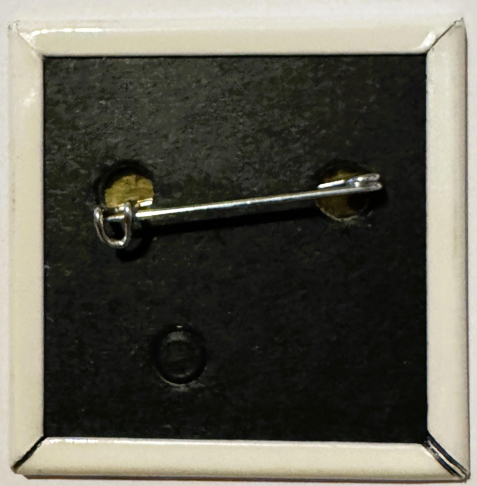 Silver pin-back button with brass fastening displayed against dark textured background in white frame, shown at angle