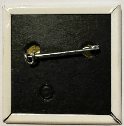 Silver pin-back button with brass fastening displayed against dark textured background in white frame, shown at angle