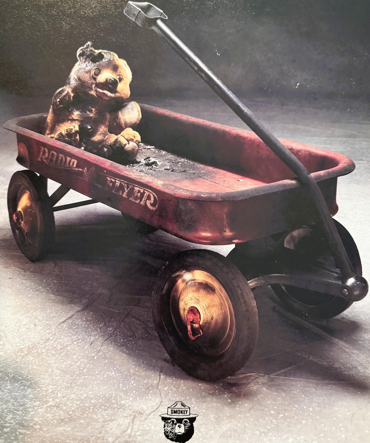 Vintage Smokey Bear poster showing charred teddy bear in red wagon with shovel, Smokey logo below, circa 1980s