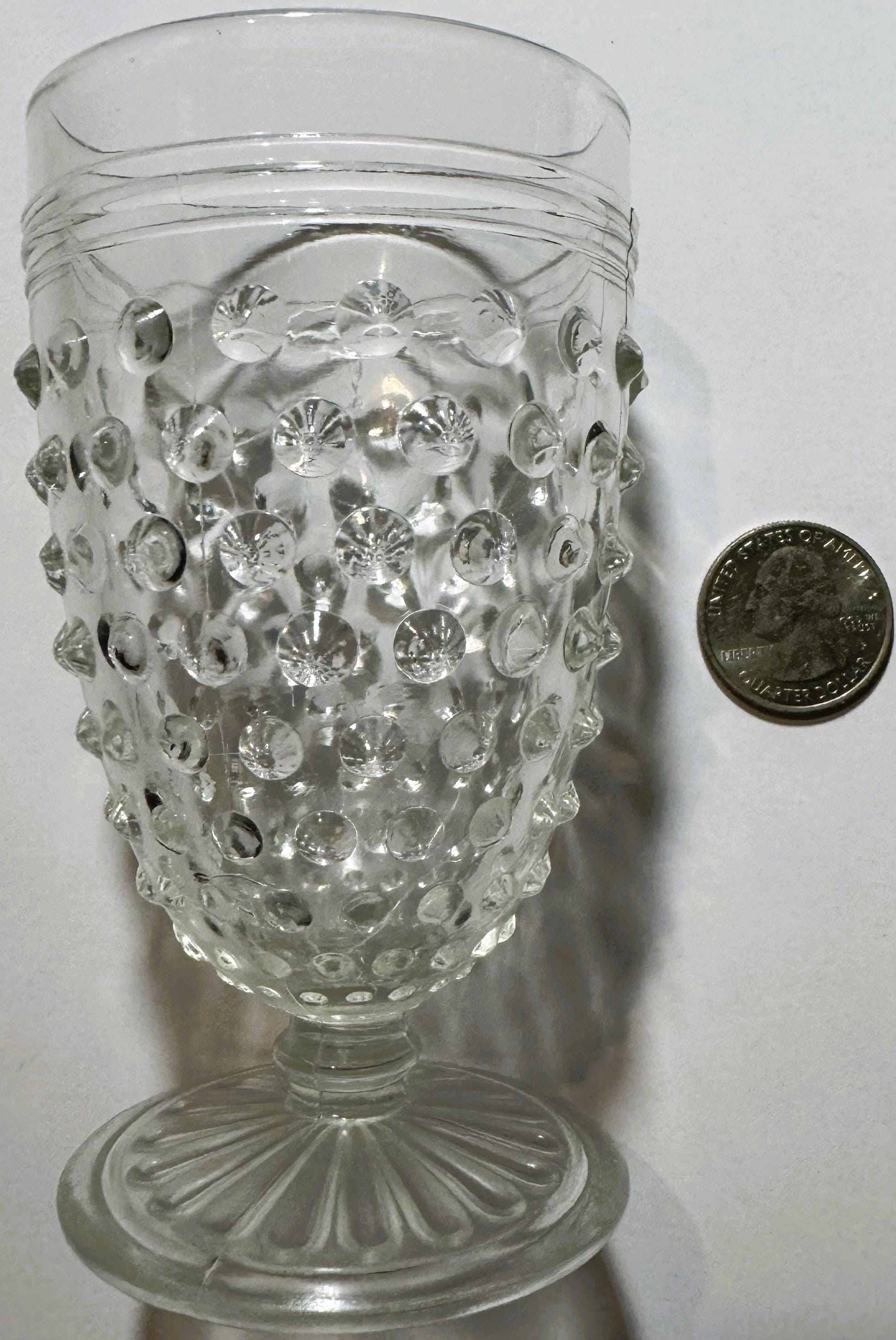 Clear glass hobnail tumbler with pedestal base next to a quarter for scale, showing dimpled surface detail and flared rim.