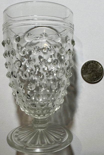 Clear glass hobnail tumbler with pedestal base next to a quarter for scale, showing dimpled surface detail and flared rim.