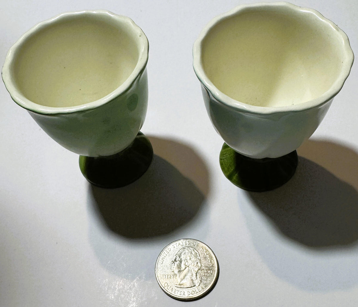Two vintage ceramic egg cups with cream interiors and green exteriors, pedestal bases, shown with a U.S. quarter for scale against white background.