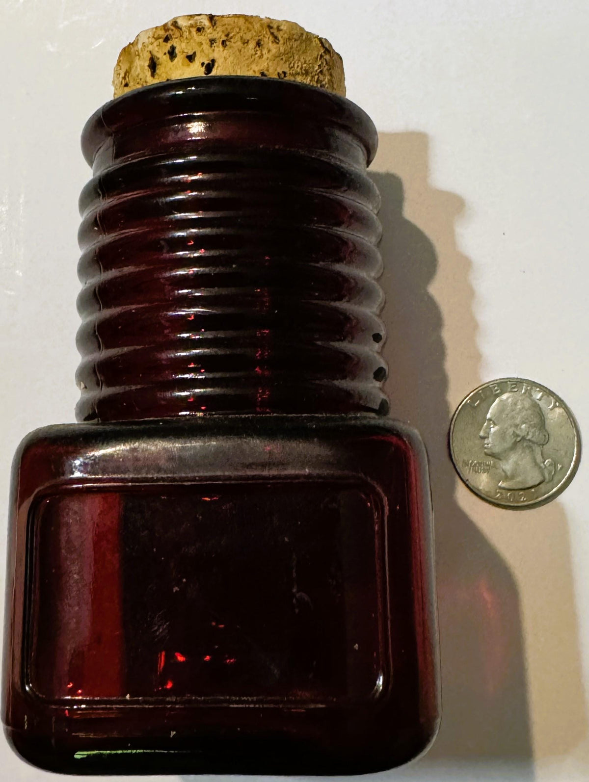Ruby red glass bottle with ribbed neck and cork stopper shown beside a US quarter for scale.