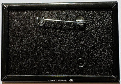 Vintage enamel pin in black packaging with metal clasp, shown from above against textured background