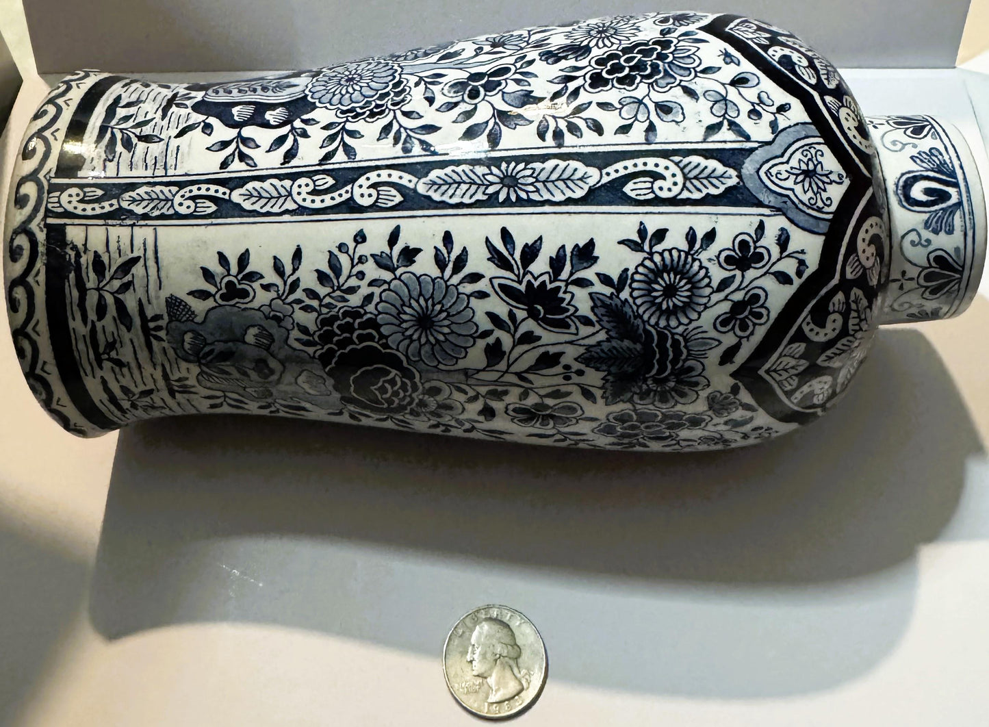 Cylindrical blue and white ceramic vase with floral patterns, shown horizontally next to a quarter for scale