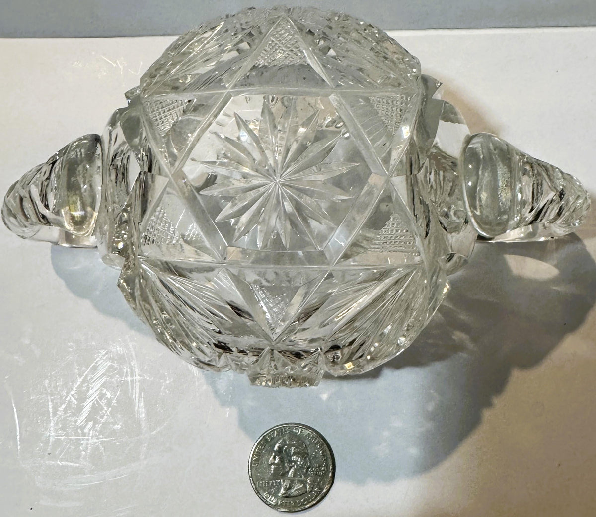 Clear cut glass two-handled sugar bowl with geometric star and fan patterns, shown from above with a quarter for scale